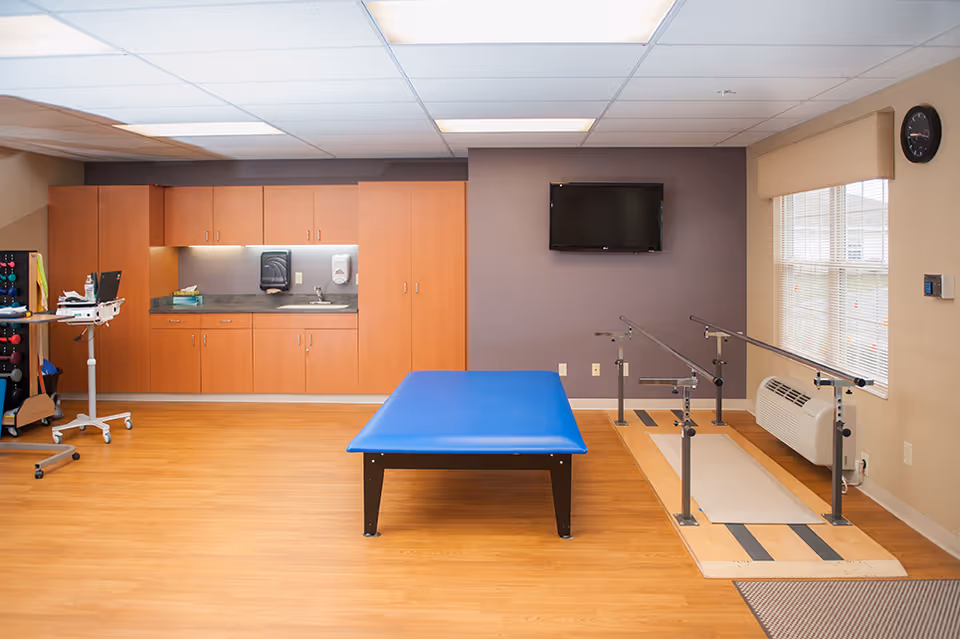 A rehabilitation or physical therapy room with a blue padded therapy table in the center, parallel bars for walking exercises near a window, wooden cabinets with a countertop and sink along the back wall, a wall-mounted TV, and medical equipment on a rolling stand to the left.
