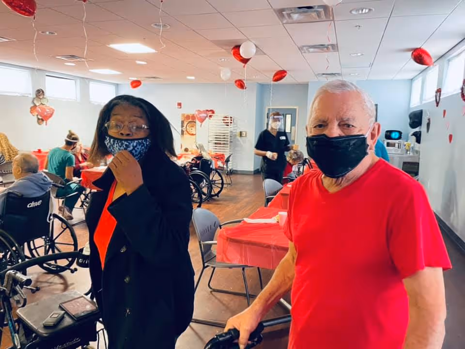 Two elderly individuals wearing face masks stand in a decorated common room with red and white heart-shaped balloons hanging from the ceiling. Several other seniors, some in wheelchairs, are seated at tables covered with red tablecloths. A staff member wearing a face shield is visible in the background.