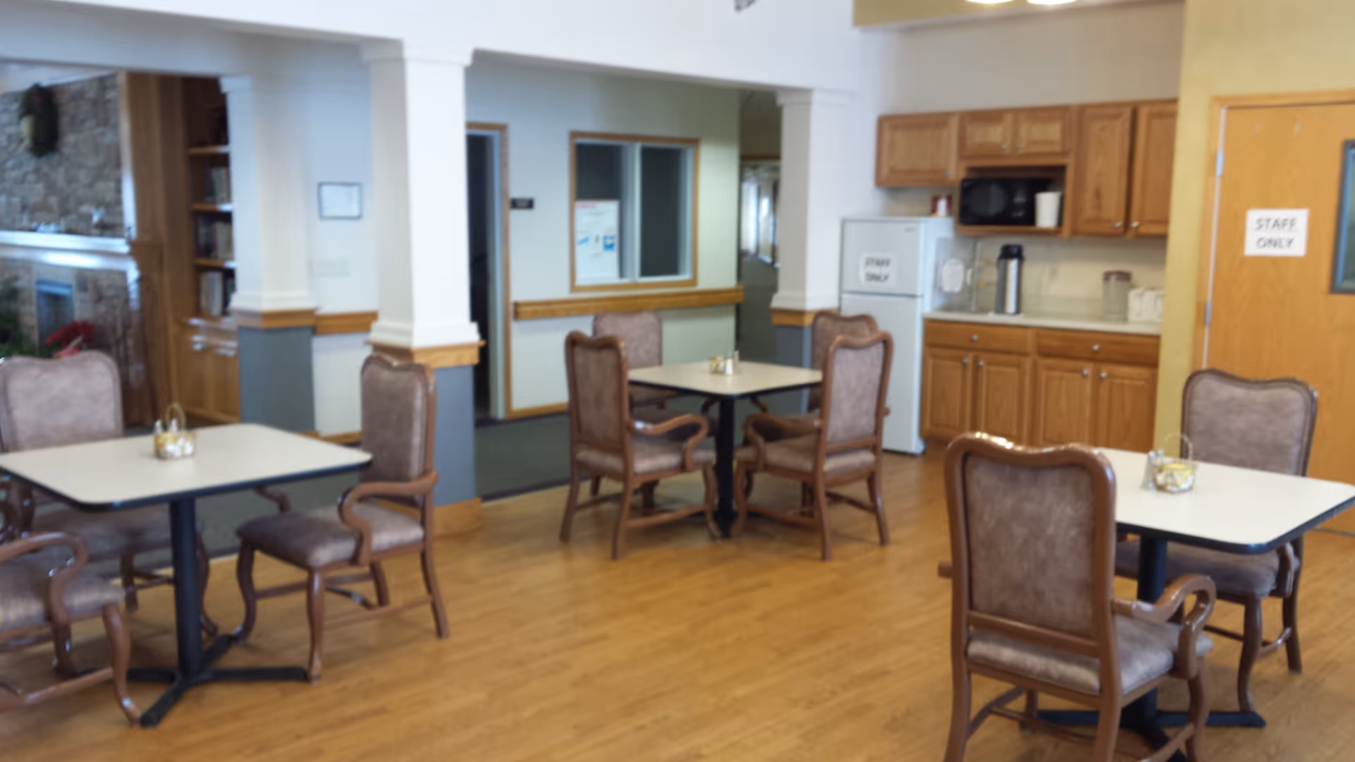 A dining area in an assisted living facility with several tables and cushioned chairs arranged on a wooden floor. In the background, there is a kitchenette with wooden cabinets, a microwave, and a refrigerator labeled 'Staff Only'. The room has white walls with wooden trim and columns, and a stone fireplace is partially visible on the left side.