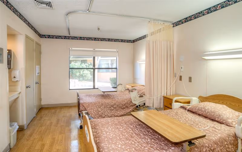 Shared patient bedroom containing three adjustable hospital beds with overbed tables, a privacy curtain, a window, and wood-look flooring.