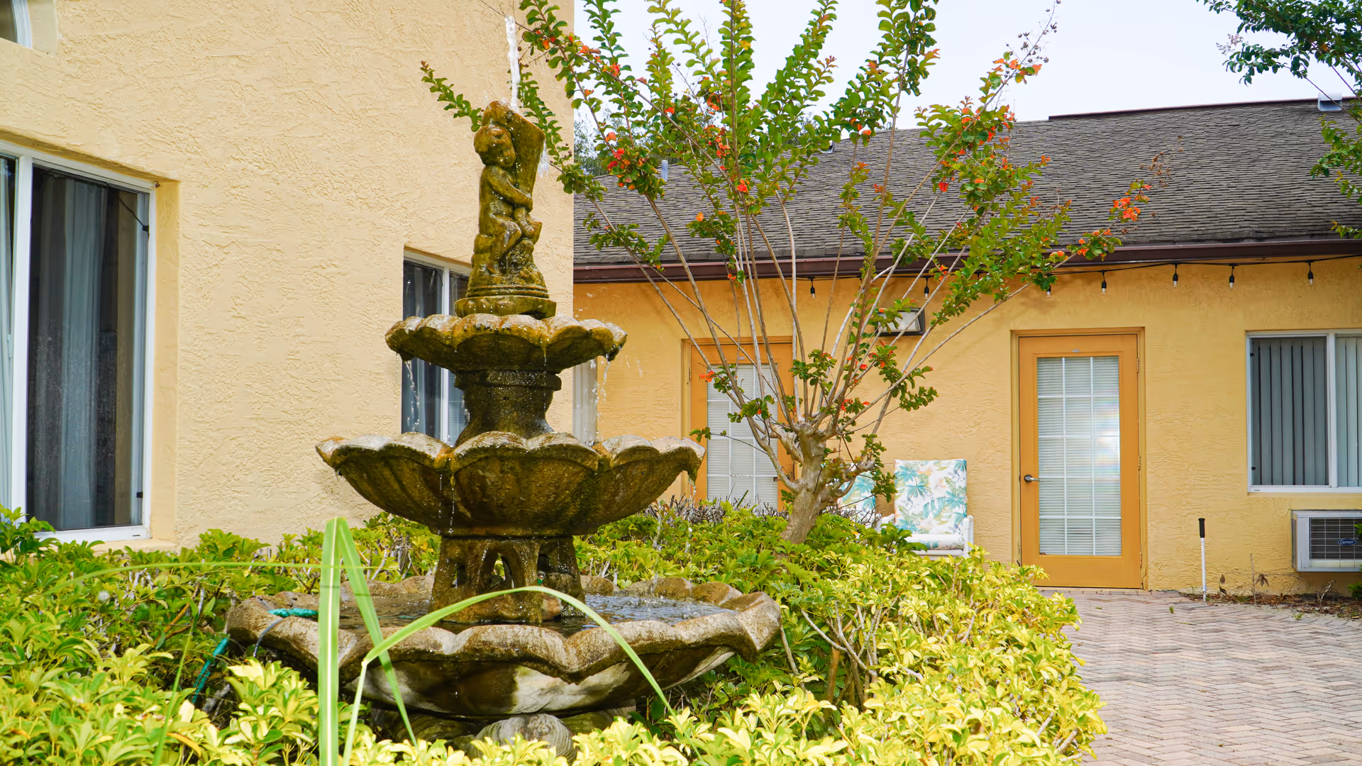 Tiered stone fountain surrounded by shrubs in a courtyard outside a yellow stucco building with doors and windows.