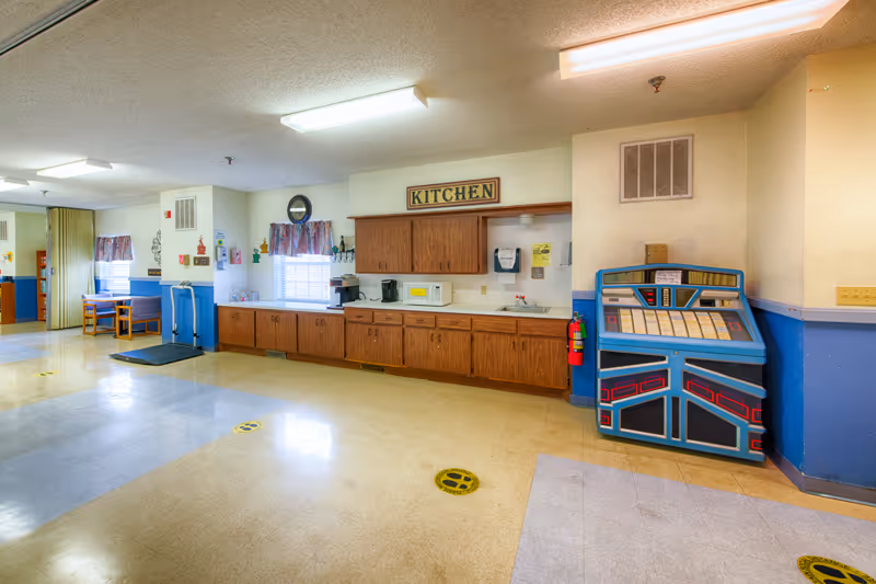 Community kitchen area with long wooden cabinets, a microwave and coffee maker beneath a 'KITCHEN' sign and a jukebox along the wall.