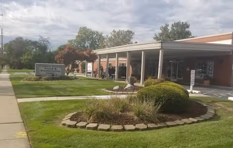 Exterior view of Mission Point Nursing & Physical Rehabilitation Center of Warren showing a brick building with a covered entrance, a landscaped circular garden with bushes and plants, a sidewalk, and a sign indicating the facility name and health & rehabilitation center.
