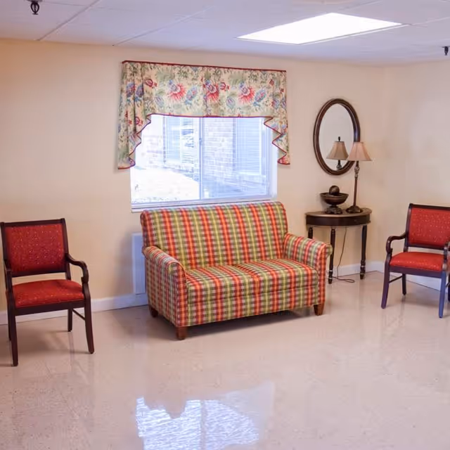A small sitting area with a plaid patterned loveseat in red, green, and yellow tones, flanked by two red cushioned wooden chairs. Behind the loveseat is a window with floral valance curtains. To the right of the loveseat is a small round wooden table with a decorative bowl and a lamp, and above the table is an oval mirror on the wall. The room has beige walls and a shiny light-colored floor.