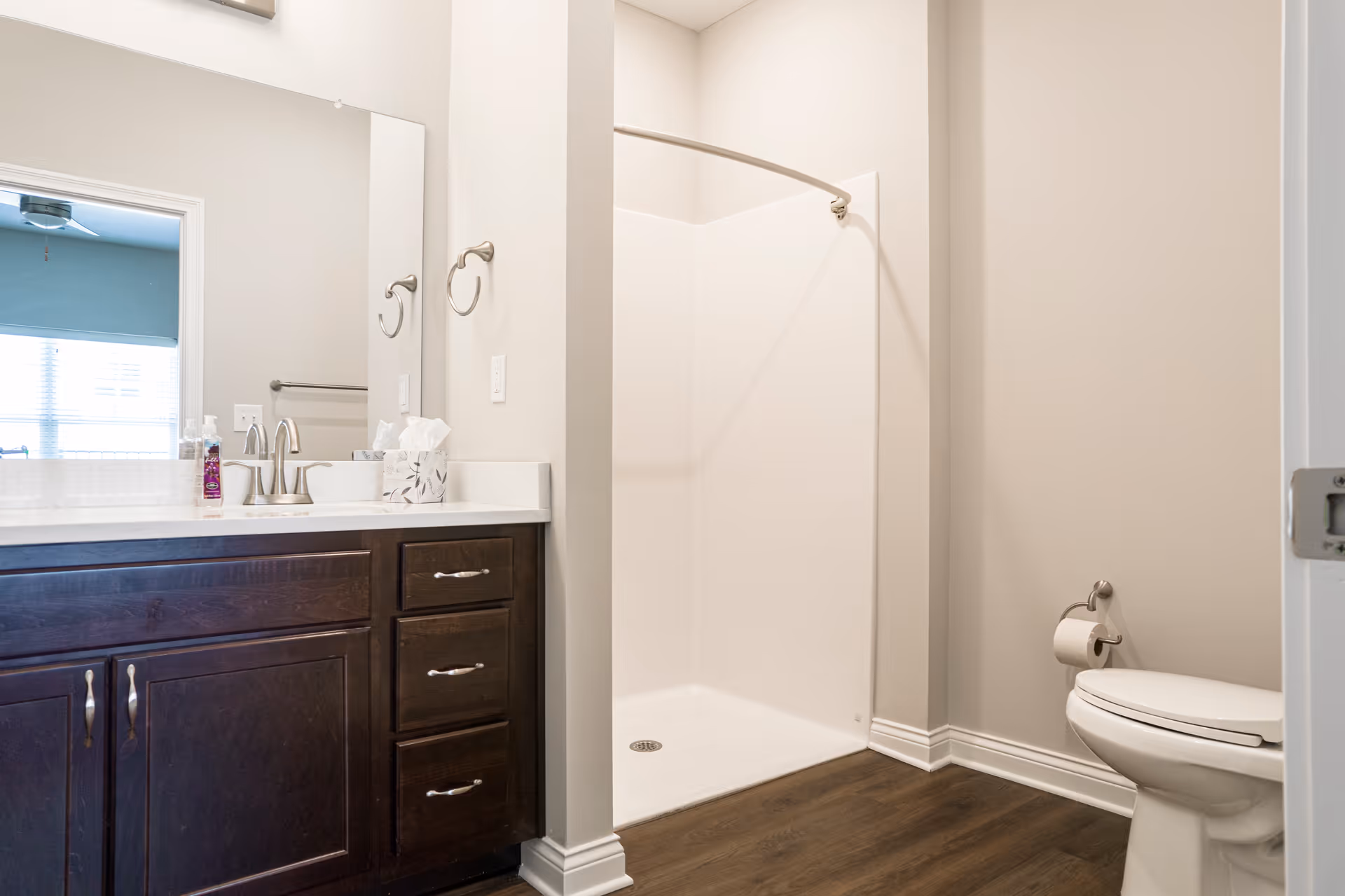 A clean bathroom featuring a dark wood vanity with a white countertop and a silver faucet. There is a large mirror above the vanity. To the right, there is a white toilet with a toilet paper holder mounted on the wall. Next to the toilet is a walk-in shower with a curved shower curtain rod. The floor is a dark wood finish and the walls are painted light gray.