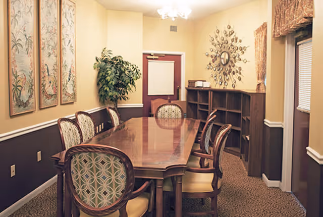 A formal dining room with a long wooden table surrounded by eight upholstered chairs. The walls are painted in two tones, with framed artwork on one side and a decorative mirror on the other. There is a potted plant in the corner and a window with a valance on the right side.