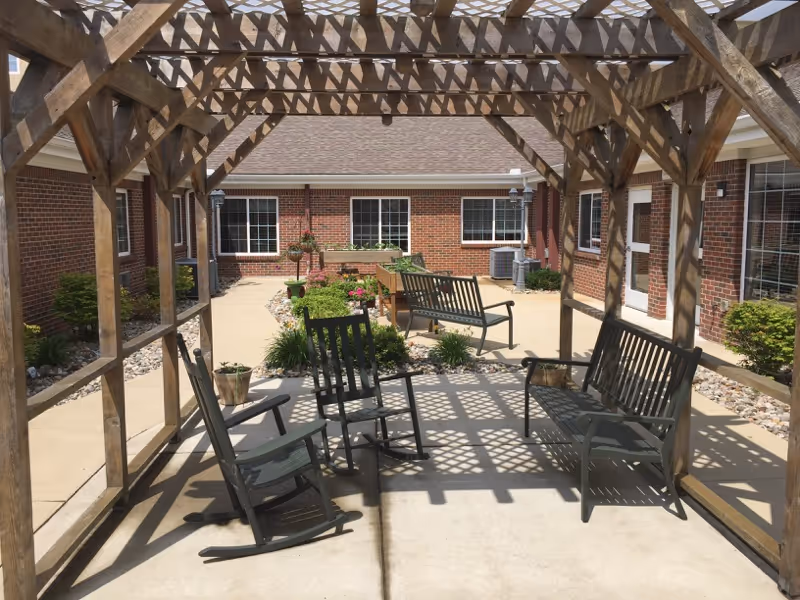 Outdoor courtyard area with a wooden pergola casting shadows on the concrete floor. Under the pergola are two rocking chairs, a bench, and a small table with a potted plant. The courtyard is surrounded by a brick building with windows and doors, and there are landscaped garden beds with plants and flowers along the edges.