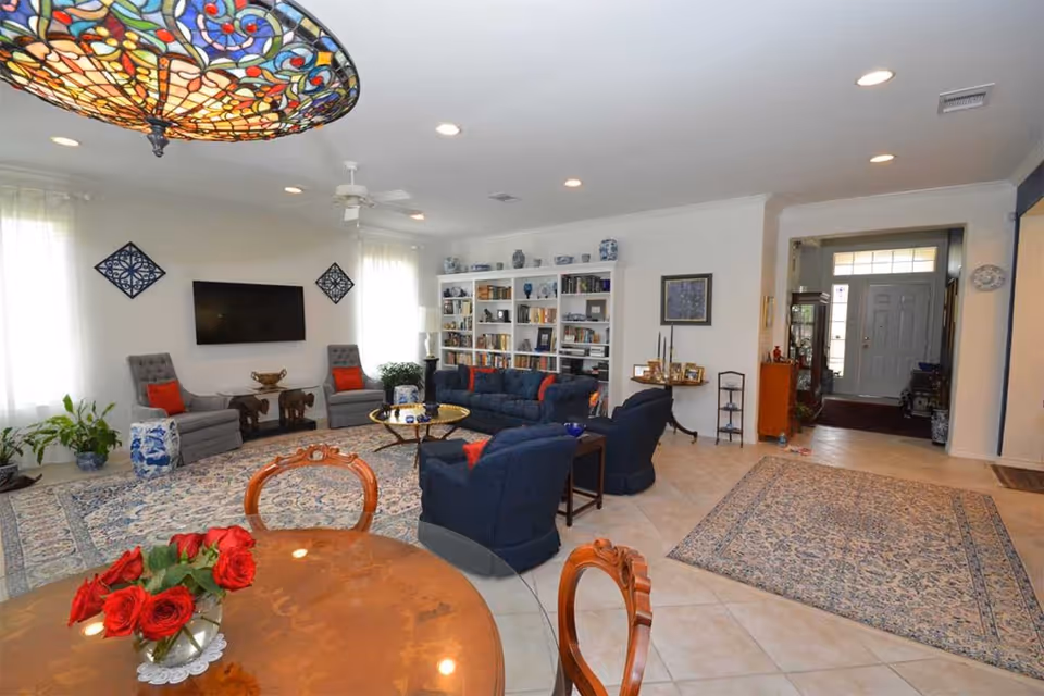 A spacious and well-lit living room with a large area rug, blue upholstered sofas and chairs, a wall-mounted flat-screen TV, a white bookshelf filled with books and decorative items, and a wooden dining table with a vase of red roses. The room has tiled flooring, white walls, and multiple windows with sheer curtains allowing natural light to enter. A colorful stained glass ceiling light fixture is partially visible.