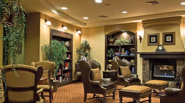 A cozy living room area with several leather armchairs and ottomans arranged around a fireplace. The room features warm lighting, built-in bookshelves with decorative items and plants, and framed artwork above the fireplace. The carpet is patterned and the walls are painted in a soft beige tone.