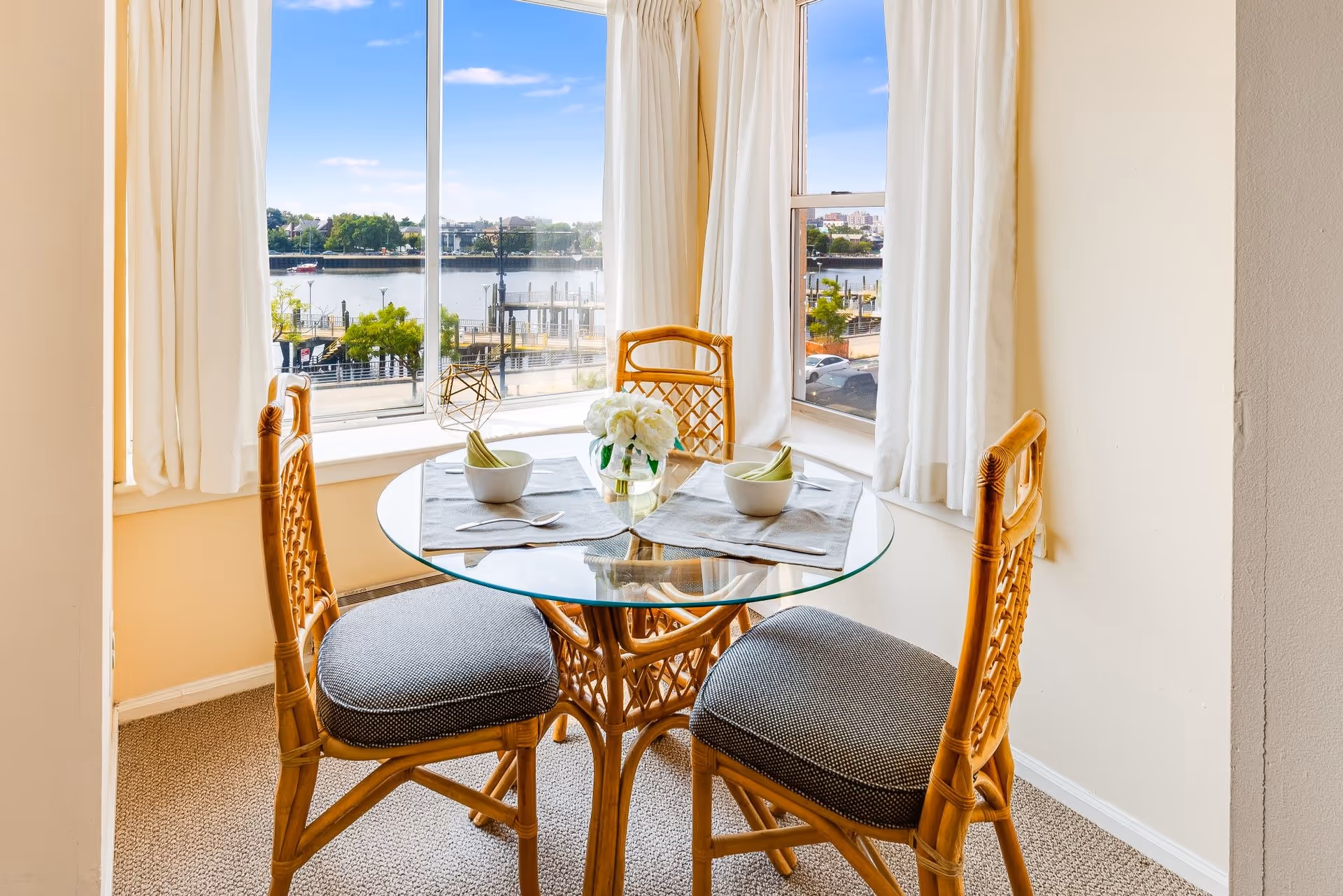 A small dining area with a round glass table and three wicker chairs with gray cushions. The table is set with two bowls, napkins, and spoons, and a small vase with white flowers is in the center. Large windows with white curtains offer a view of a waterfront and buildings across the water under a blue sky.