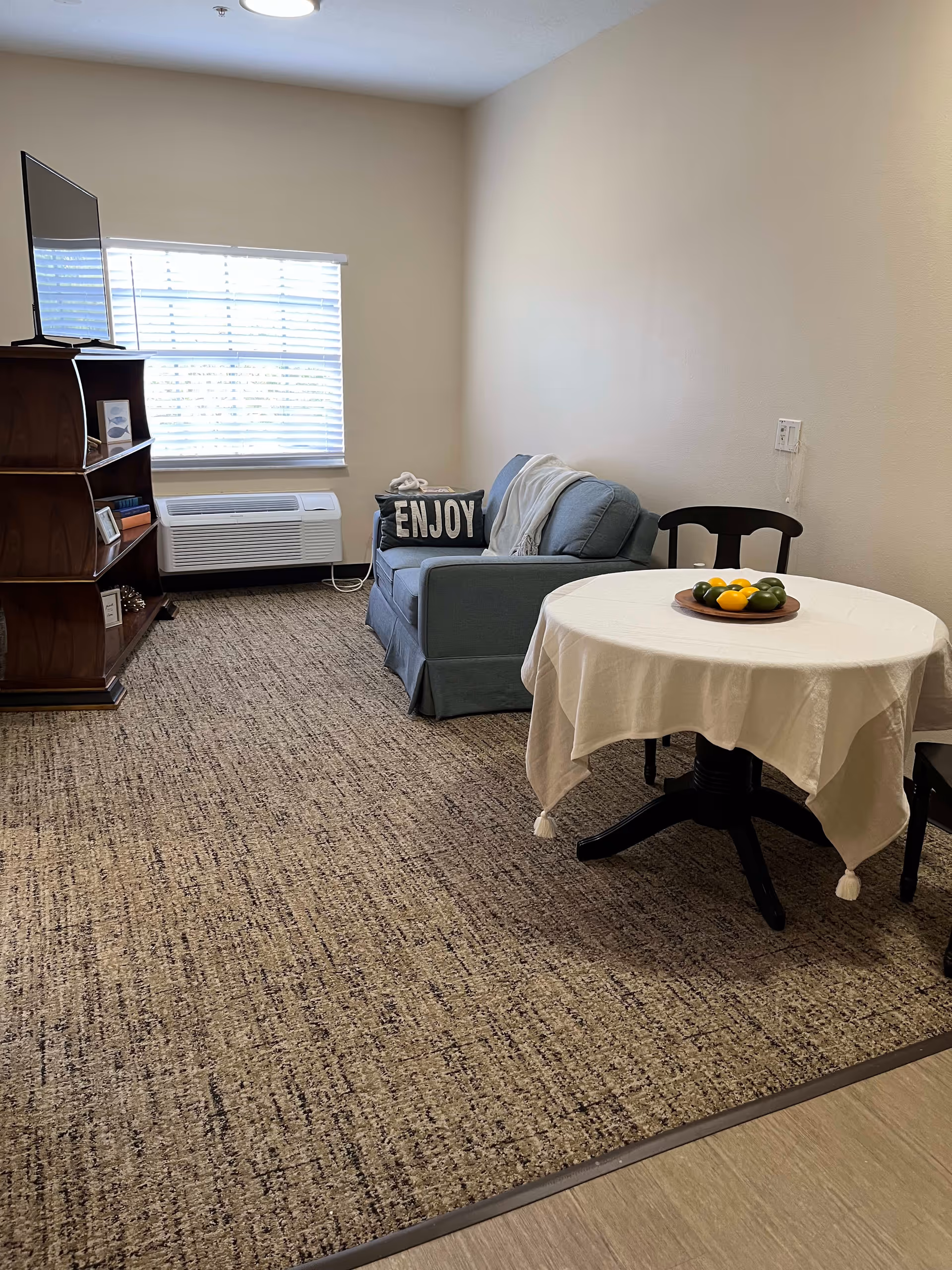 A cozy living room area with a blue loveseat that has a pillow with the word 'ENJOY' on it. There is a round table covered with a white tablecloth and a plate of decorative fruit on top. A wooden shelving unit holds a flat-screen TV and some books. A window with blinds and an air conditioning unit below it is on the far wall. The room has beige walls and carpeted flooring.