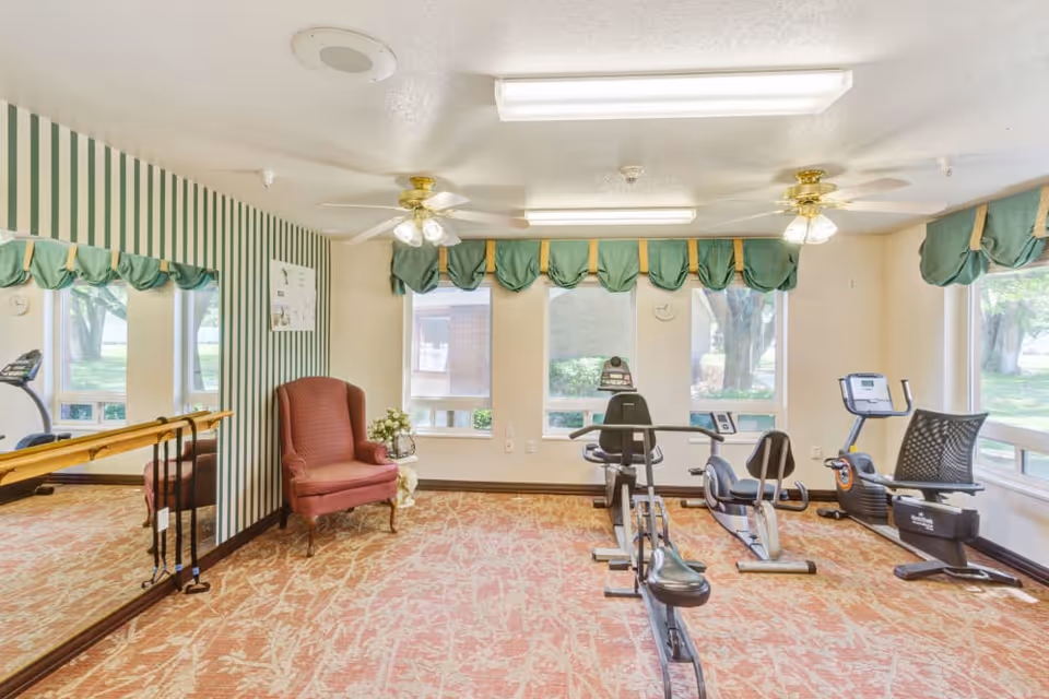 A small exercise room with three stationary bikes, a red upholstered armchair, a side table with flowers, large windows with green valances, striped wallpaper on one wall, a large mirror, and ceiling fans with lights.