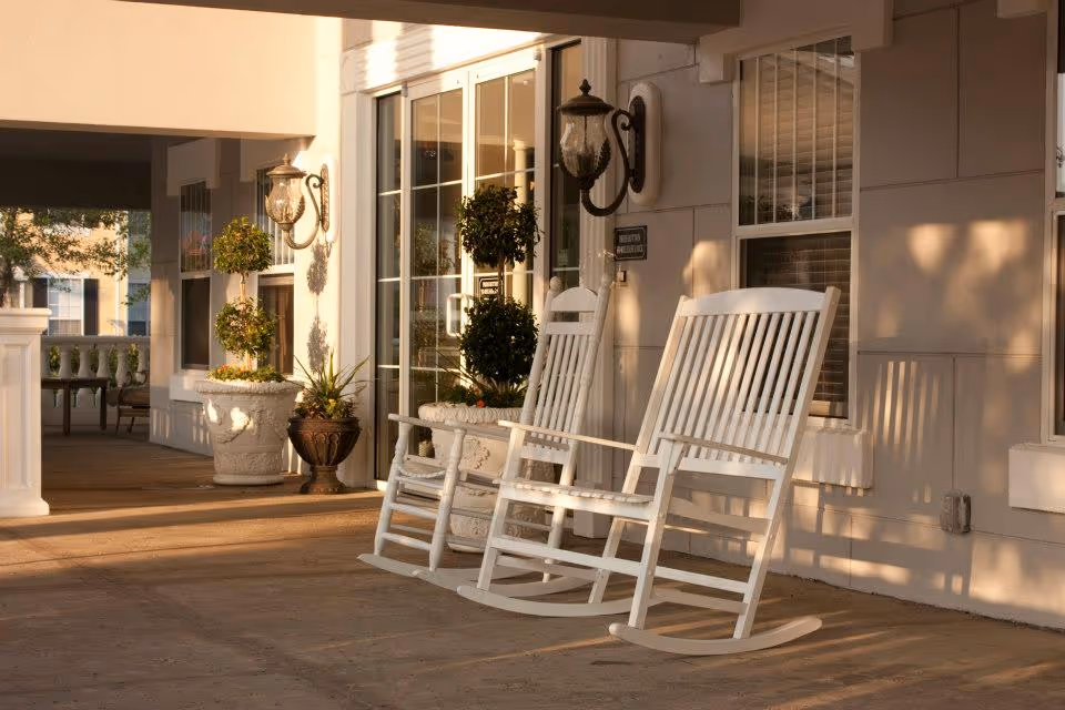 Two white wooden rocking chairs on a covered porch outside a building with large windows and decorative potted plants, bathed in warm sunlight.