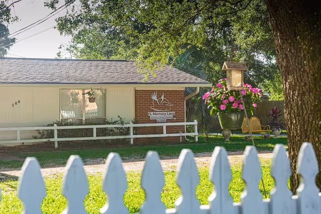 Exterior view of a single-story building with a white picket fence in the foreground. The building has a brick and light-colored siding facade with a sign that reads 'FL Memory Lane Assisted Living Facility'. There is a hanging flower pot with pink flowers and a tree on the right side of the image.