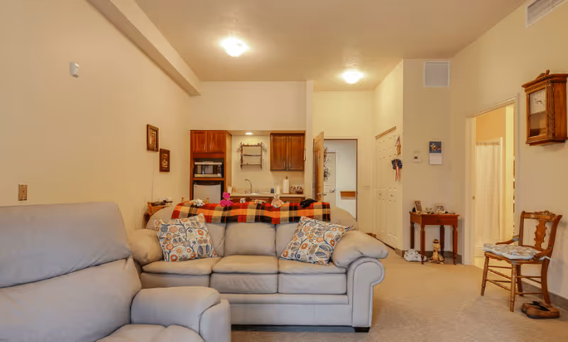 Interior view of a living area in an assisted living facility showing a beige leather sofa with patterned cushions and a matching armchair. Behind the sofa is a small kitchen area with wooden cabinets, a microwave, and a sink. The room has beige walls and carpet, with a small table and chair near a doorway leading to another room. A wall clock and framed pictures are visible on the walls.
