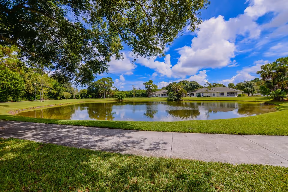 A peaceful outdoor scene at Lake Forest Park featuring a small pond surrounded by well-maintained grass and trees. A concrete pathway runs along the edge of the pond, and residential buildings are visible in the background under a partly cloudy blue sky.
