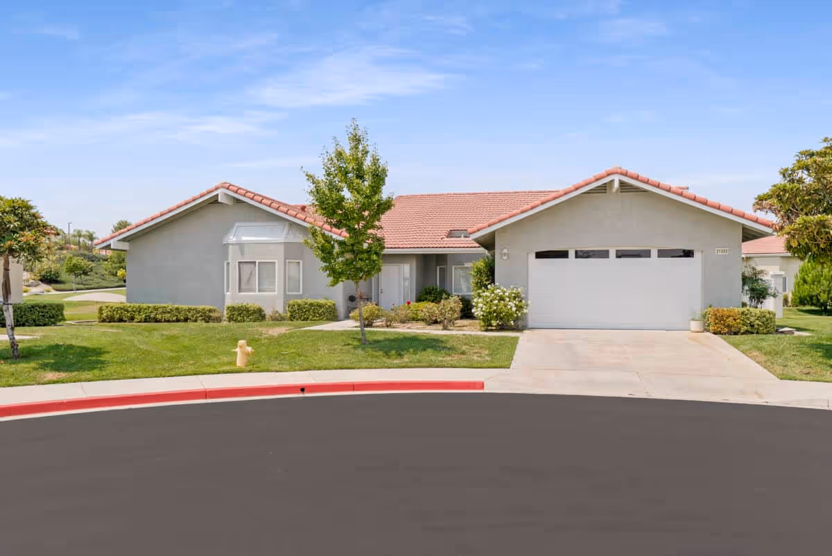 Single-story residential building with a light gray exterior and red-tiled roof, featuring a garage on the right side and a small tree and shrubs in front, under a clear blue sky.