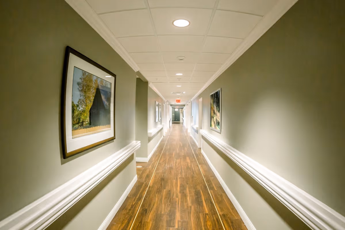 A long, well-lit hallway with wooden flooring and light green walls. The hallway has white trim and handrails on both sides, with framed pictures hanging on the walls. The ceiling has recessed lighting and a visible exit sign at the far end.
