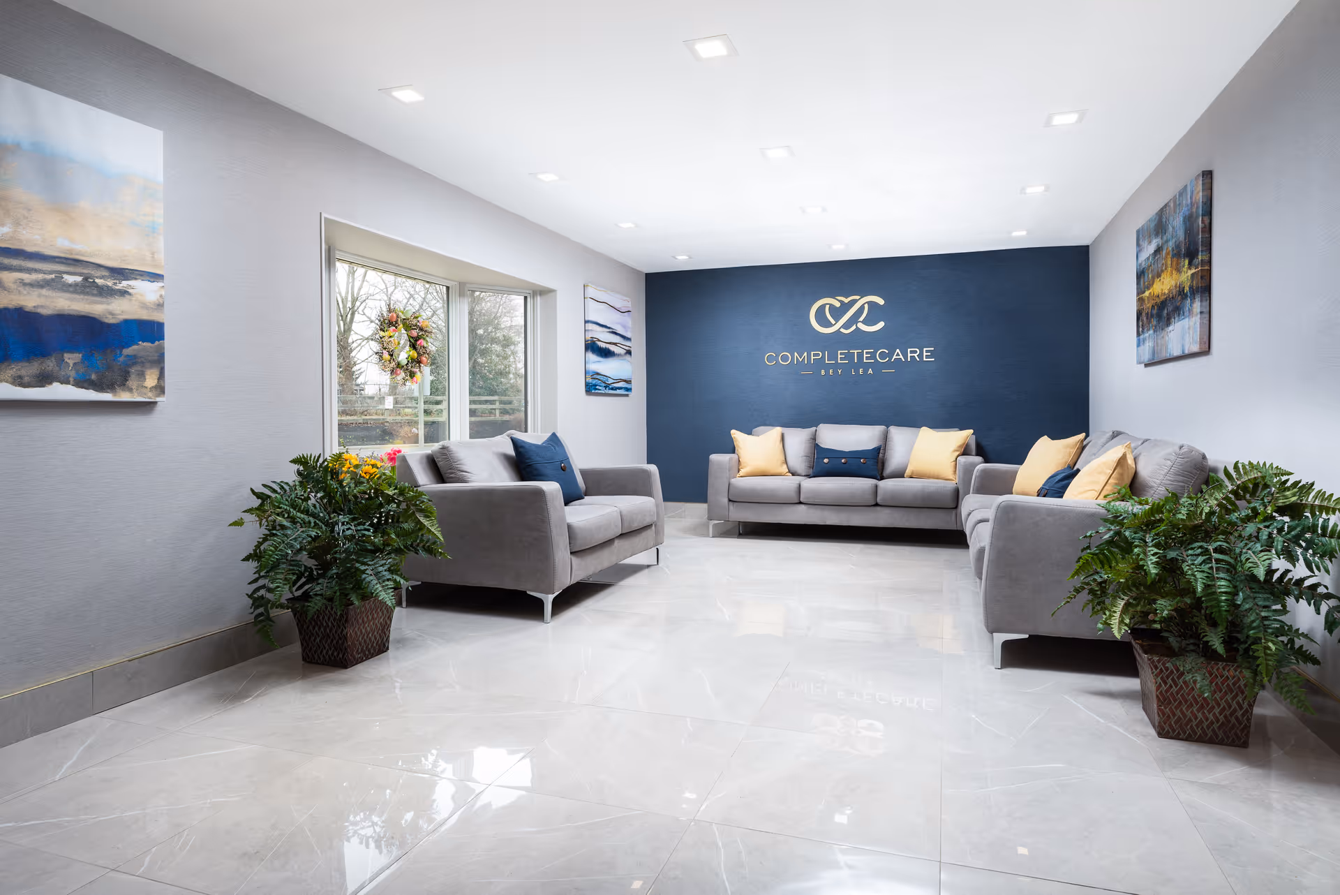 A bright and modern lounge area with three gray sofas arranged around the room, decorated with yellow and blue cushions. The back wall is painted dark blue and features the Complete Care at Bey Lea logo in gold. There are large windows on one side letting in natural light, two large green potted plants, and abstract paintings on the walls.