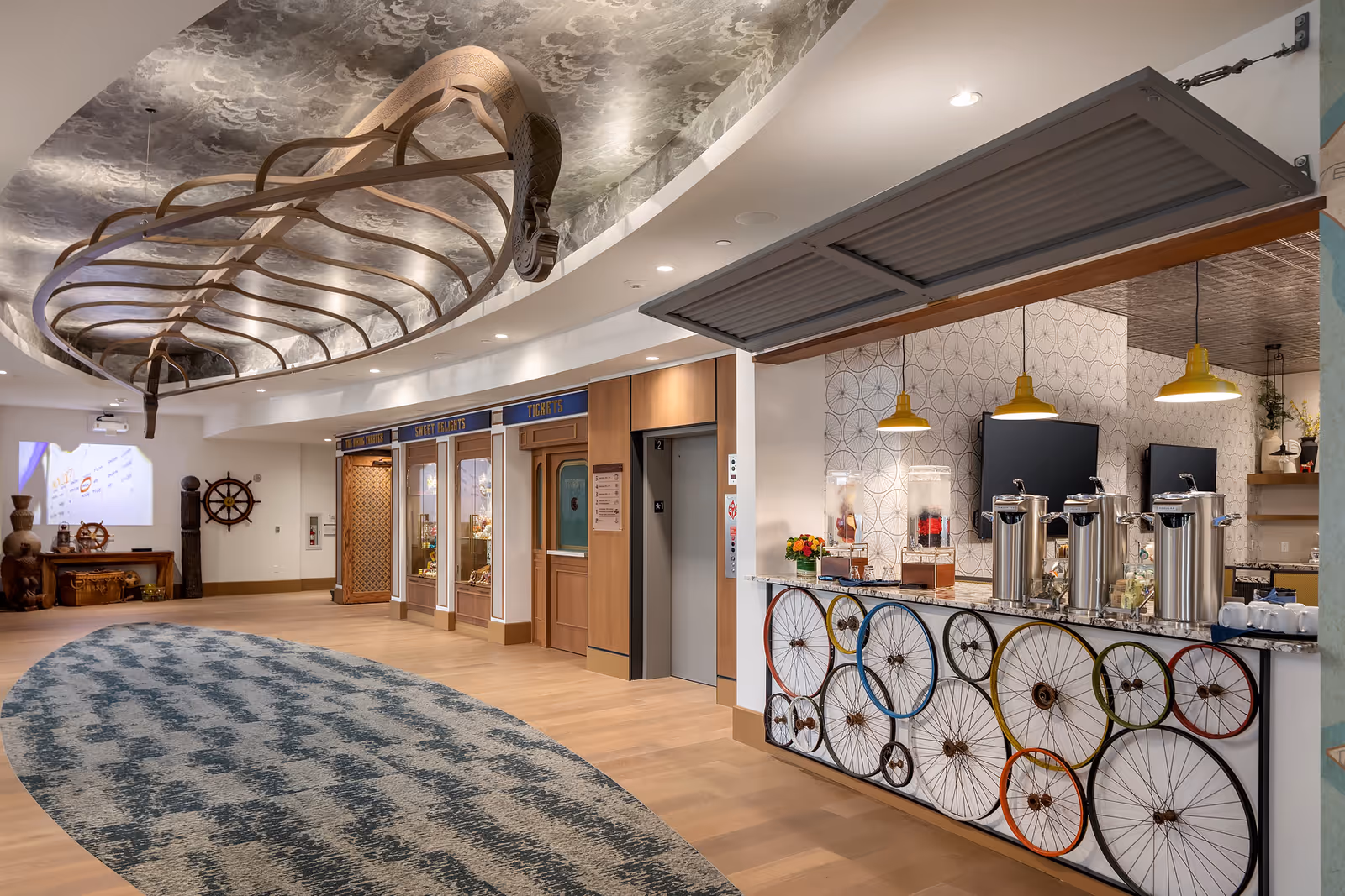 Interior view of a senior living facility hallway with a decorative ceiling featuring a wooden boat frame. On the right side, there is a refreshment counter decorated with colorful bicycle wheels and equipped with beverage dispensers and cups. The back wall has an elevator and display windows labeled 'Tickets' and 'Sweet Delights'. The floor has a patterned carpet runner, and the space is well-lit with recessed lighting and pendant lamps.