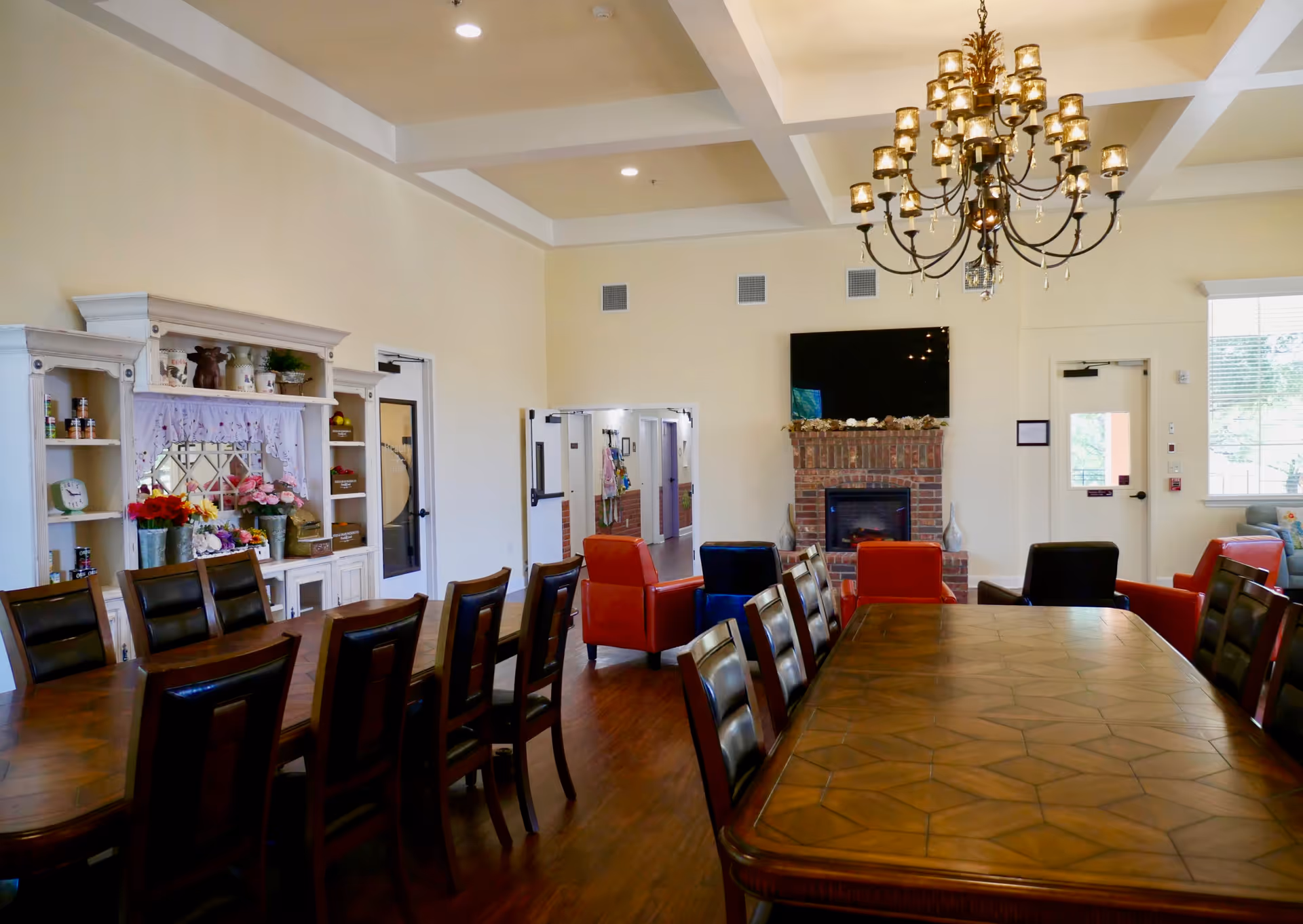 A spacious dining room with two long wooden tables surrounded by chairs. In the background, there is a brick fireplace with a flat-screen TV mounted above it. Several colorful armchairs are arranged near the fireplace. A large chandelier hangs from the ceiling. There is a white cabinet with shelves holding decorative items and flowers on the left side, and windows letting in natural light on the right.