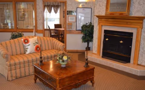 A cozy living room area featuring a striped sofa with floral pillows, a wooden coffee table with a floral centerpiece and two candlesticks, a fireplace with a wooden mantel, and a potted plant near a doorway leading to a dining area with a table and chairs.