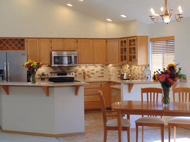 A bright kitchen and dining area with wooden cabinets, a stainless steel refrigerator and microwave, a tiled backsplash, and a countertop with flowers in vases. The dining table has wooden chairs and a vase with colorful flowers. A window with wooden blinds allows natural light into the room.