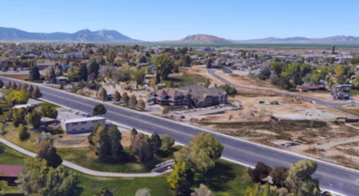 Aerial view of a suburban area with a main road, scattered buildings, trees, and mountains in the distance.