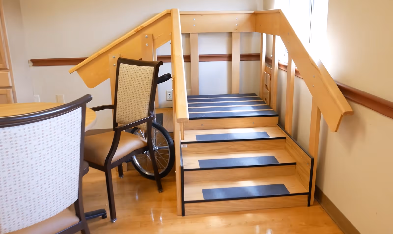 A small wooden staircase with handrails on both sides inside a room, next to a round table with chairs and a wheelchair partially visible behind one chair.