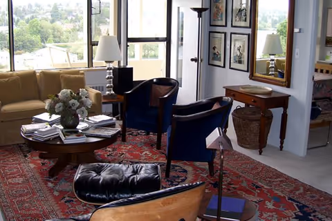 A cozy living room with large windows showing a green outdoor view. The room features a beige sofa, two dark blue armchairs, a black leather lounge chair, a round wooden coffee table with a flower vase and magazines, a red patterned rug, a wooden side table with a lamp, framed artwork on the wall, a large mirror, and a woven basket under a small wooden console table.