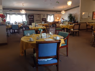 A spacious dining room with multiple round tables covered with yellow tablecloths, each set with cups, saucers, and small flower arrangements. The room has large windows with curtains, chandeliers hanging from the ceiling, and several chairs around each table. The overall atmosphere is warm and inviting.