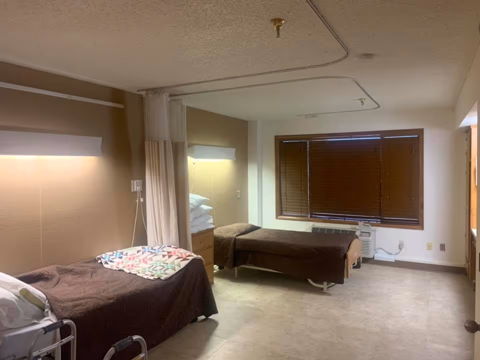 A room with two single beds covered with brown blankets, separated by a curtain. Each bed has a small bedside table with stacked pillows and a wall-mounted light above. The room has a large window with closed wooden blinds and a beige and white color scheme.