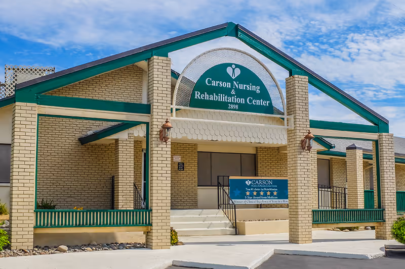 Front entrance of Carson Nursing & Rehabilitation Center with brick columns, green-trimmed portico, sign, and steps.