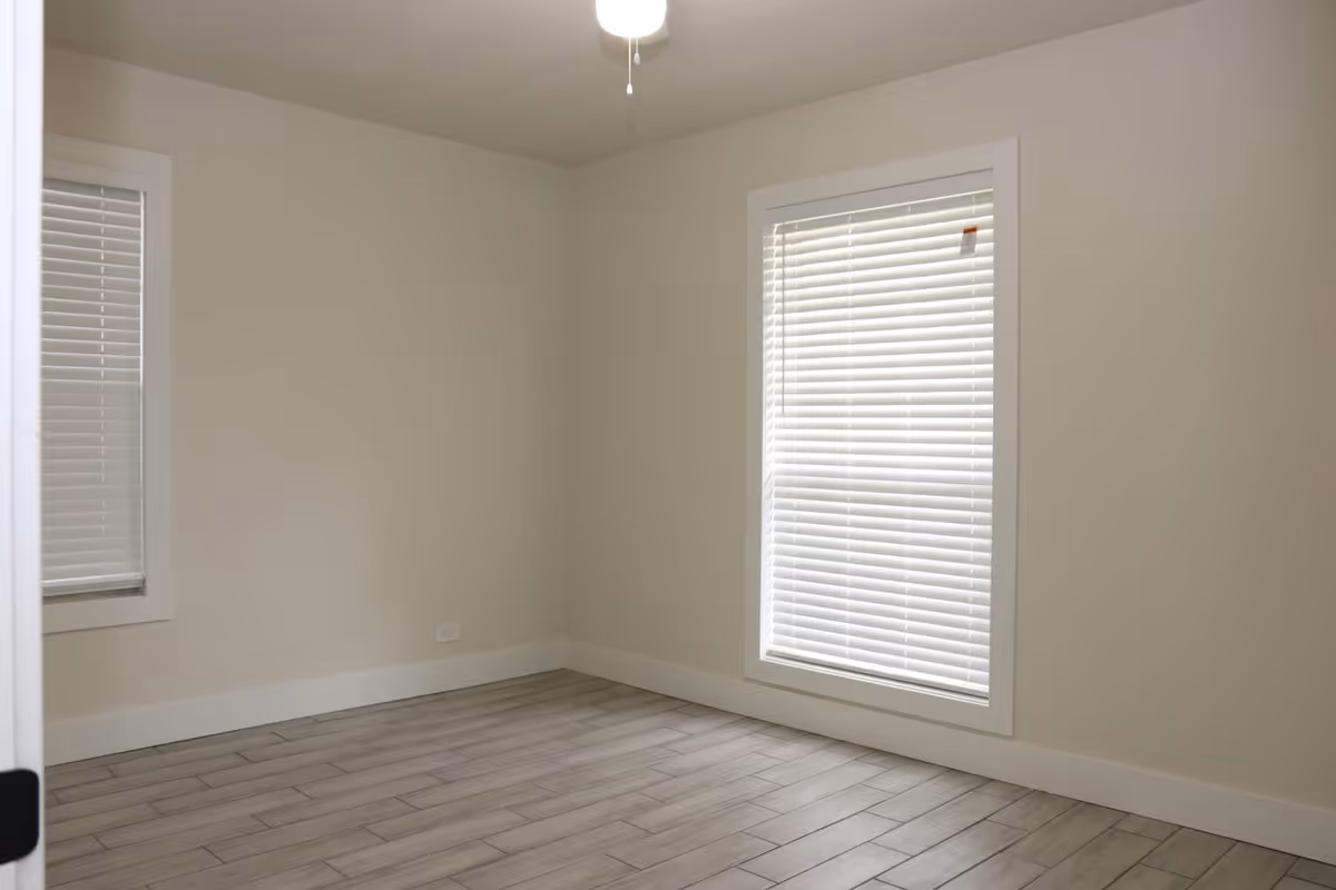 Empty room with light-colored walls, two windows with white blinds, a ceiling fan with light, and wood-look tile flooring.
