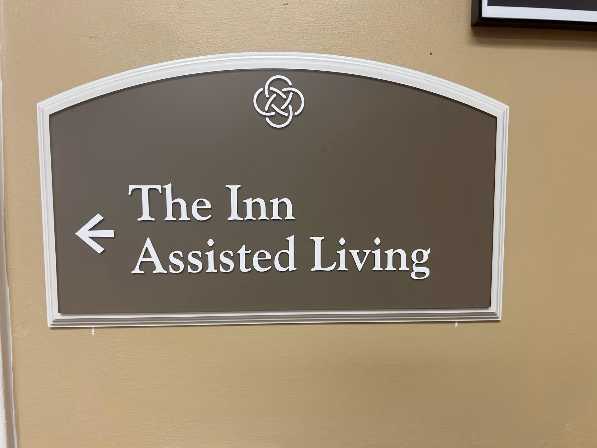 A wall-mounted sign with a brown background and white border and text. The sign reads 'The Inn Assisted Living' with an arrow pointing to the left. There is a decorative emblem above the text.