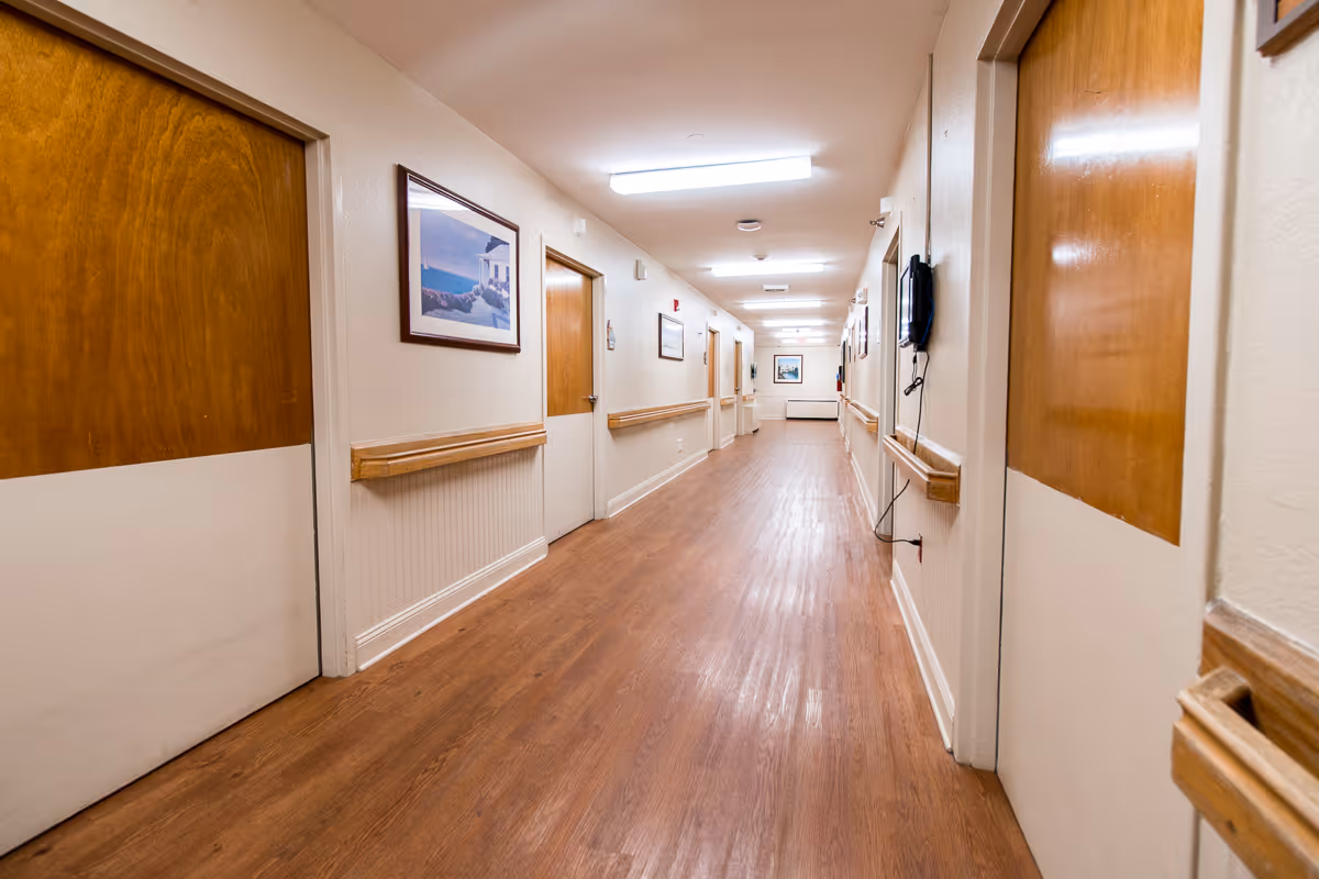 Well-lit long hallway in a care facility with wood doors, handrails, framed artwork and a wooden floor.
