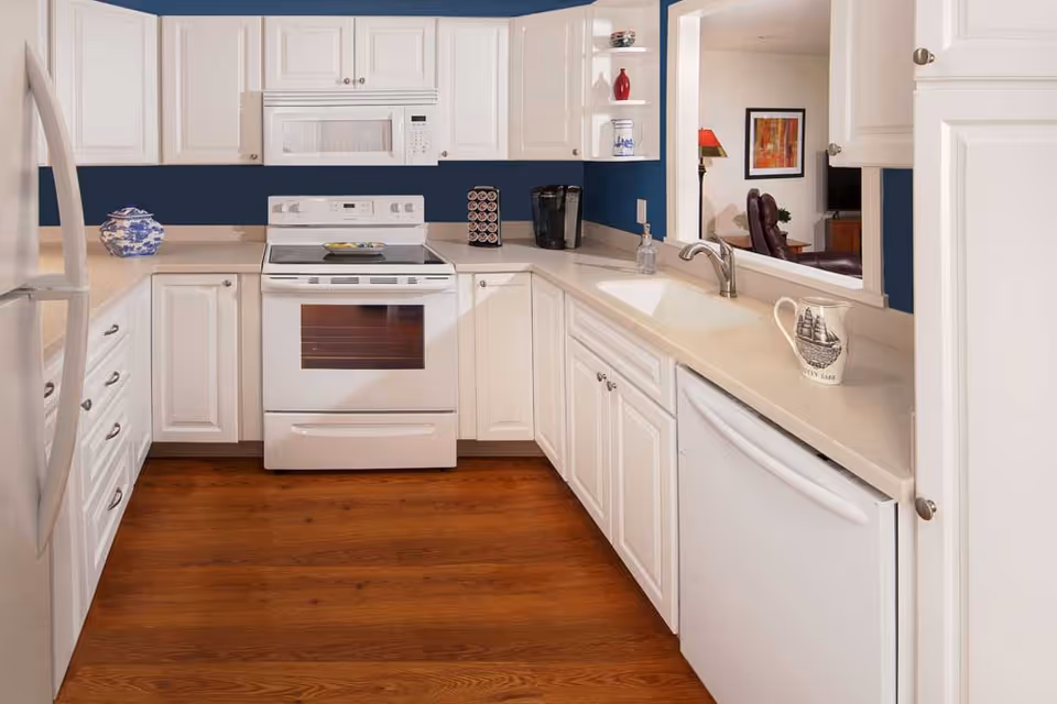 A clean, modern kitchen with white cabinets, a white stove with oven, a white microwave above it, a white dishwasher, and a white refrigerator. The countertops are light-colored, and the floor is wooden. There are decorative items on the counters including a blue and white ceramic jar, a coffee maker, a soap dispenser, and a mug with a ship design. A pass-through window looks into a living room area with a lamp, framed artwork, and a TV.