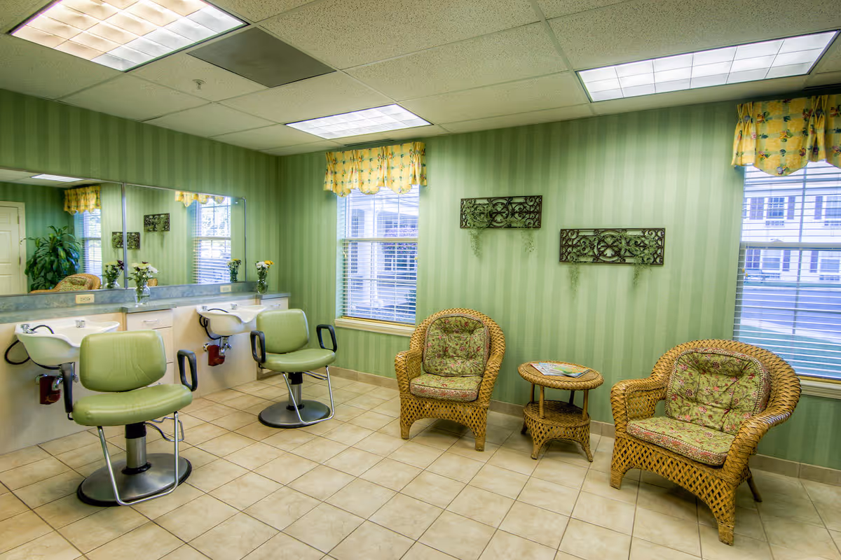 A bright salon/lounge with green styling chairs and wash basins on the left and wicker armchairs with a small table on the right against green striped walls.