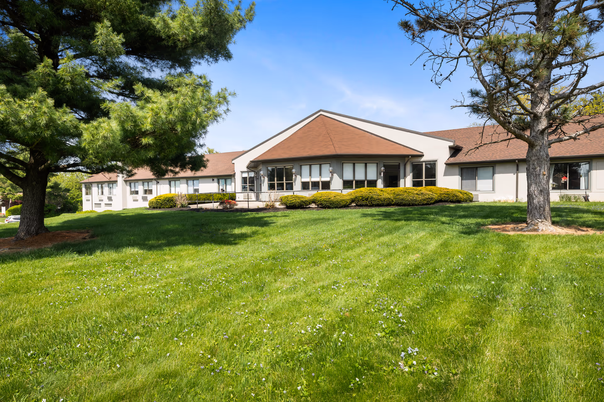 Exterior view of Willow Brook Rehabilitation and Healthcare Center showing a single-story building with a brown roof, surrounded by green grass and trees under a clear blue sky.