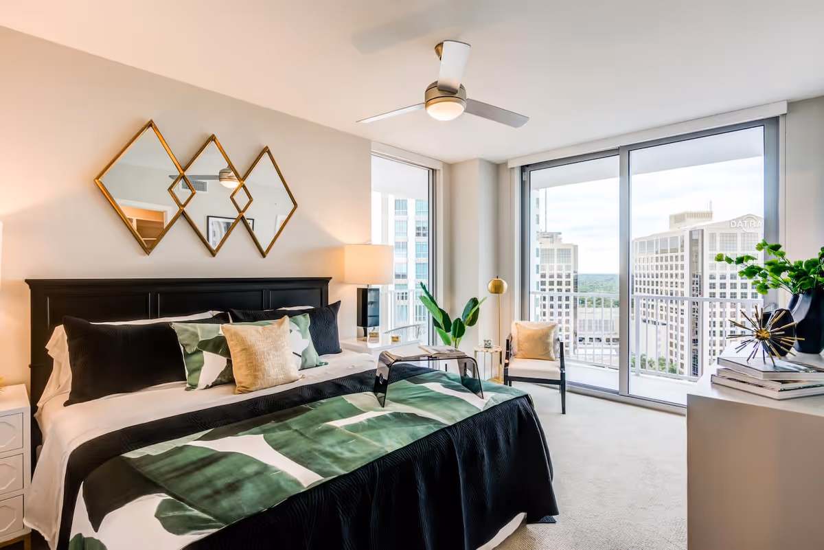 A modern bedroom with a large bed featuring black and green leaf-patterned bedding, decorative pillows, and a black headboard. Above the bed are three diamond-shaped mirrors. There is a nightstand with a lamp on one side, a ceiling fan with a light, a chair with a beige cushion near large floor-to-ceiling windows that offer a cityscape view, and a white dresser with books and a plant on top.