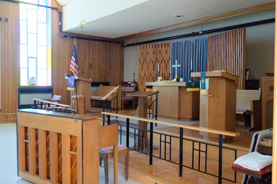 Wood-paneled chapel interior with altar, pulpit, pews, an American flag, and a stained glass window.