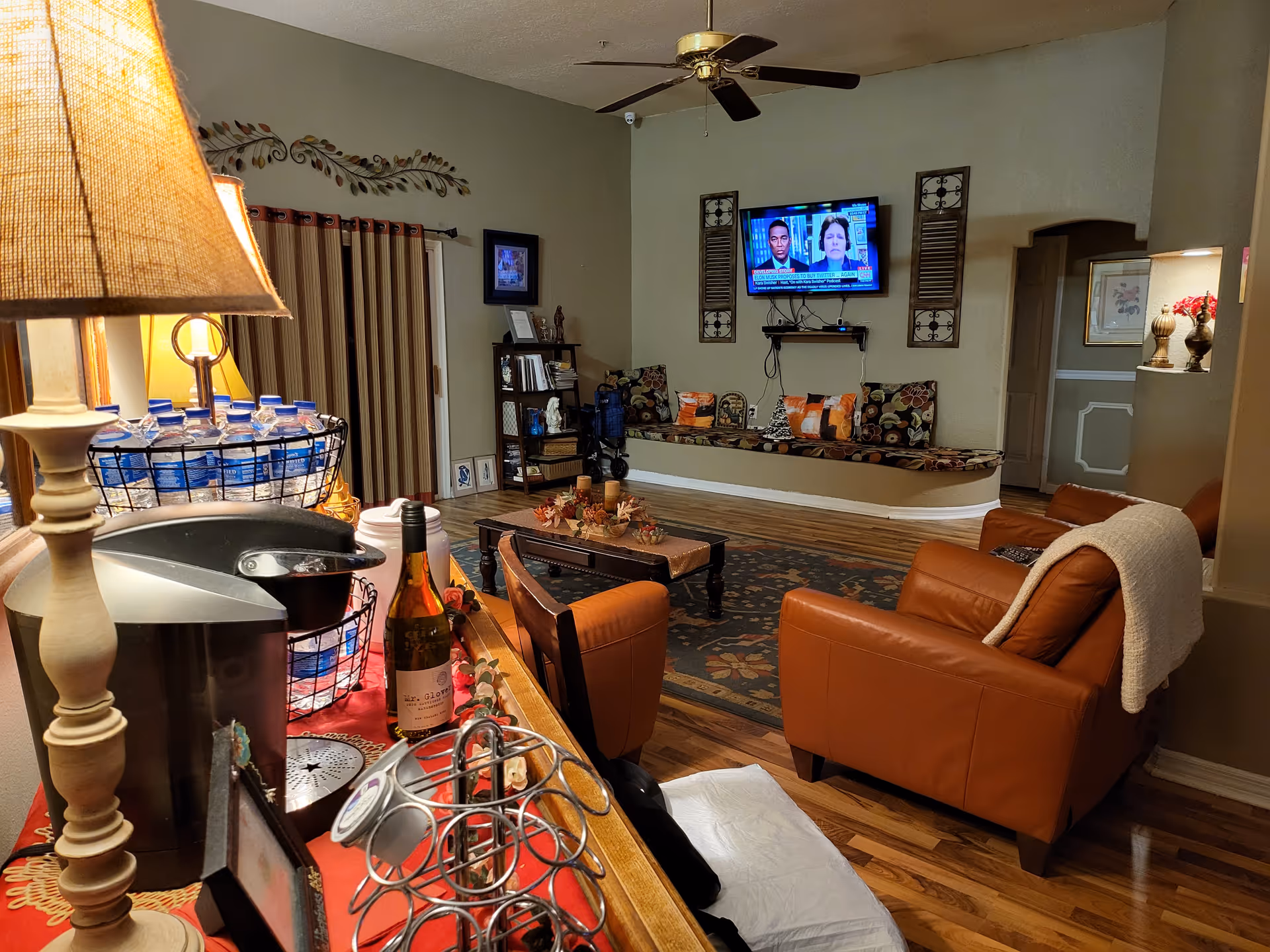 A cozy living room area with a brown leather armchair draped with a white blanket, a wooden coffee table with fall-themed decorations, and a built-in bench with colorful cushions beneath a wall-mounted TV. In the foreground, there is a table with a lamp, a coffee maker, bottled water, and a bottle of wine. The room has wooden flooring and warm lighting.