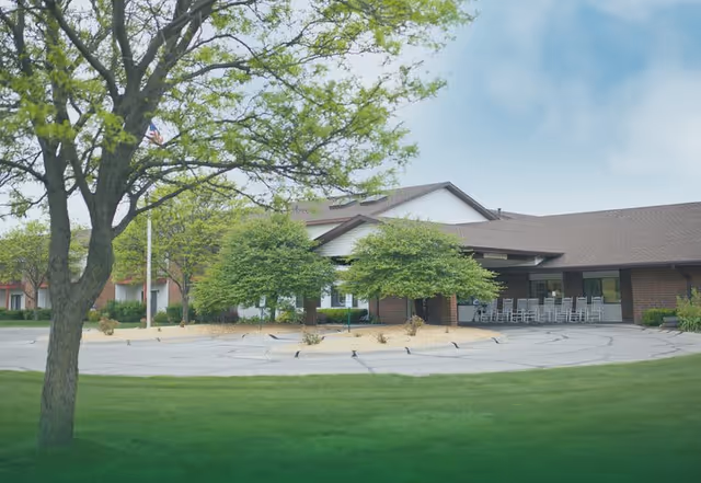 Exterior view of The Meadows senior living facility showing a single-story building with a covered entrance, several trees, a flagpole with an American flag, and a circular driveway surrounded by green grass.