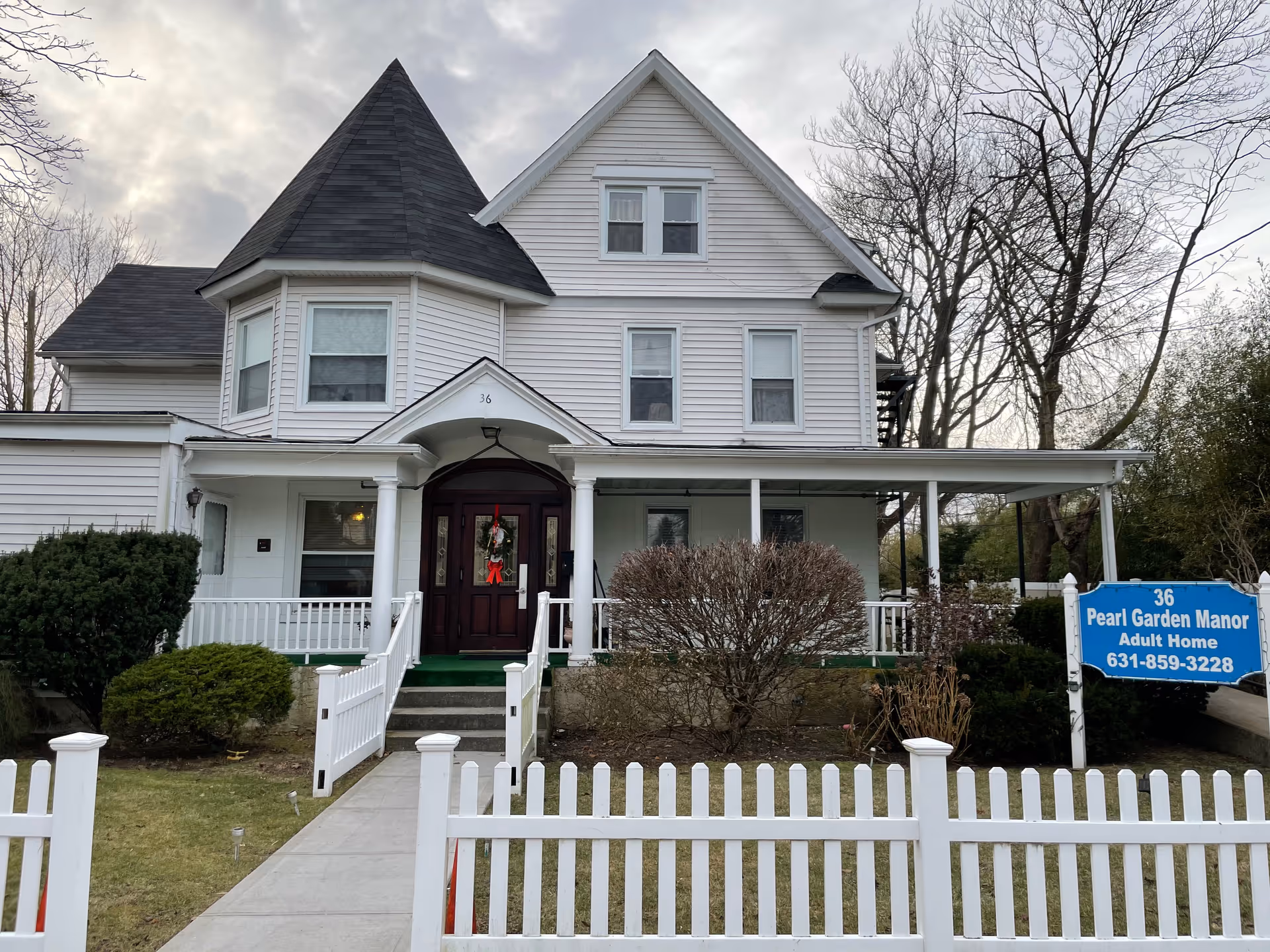 Front exterior view of a white two-story house with a pointed turret and a covered porch. There is a white picket fence in front and a blue sign on the right side that reads '36 Pearl Garden Manor Adult Home 631-859-3228'. The house has several windows and a dark wooden front door with a wreath. Leafless trees and bushes surround the property under a cloudy sky.