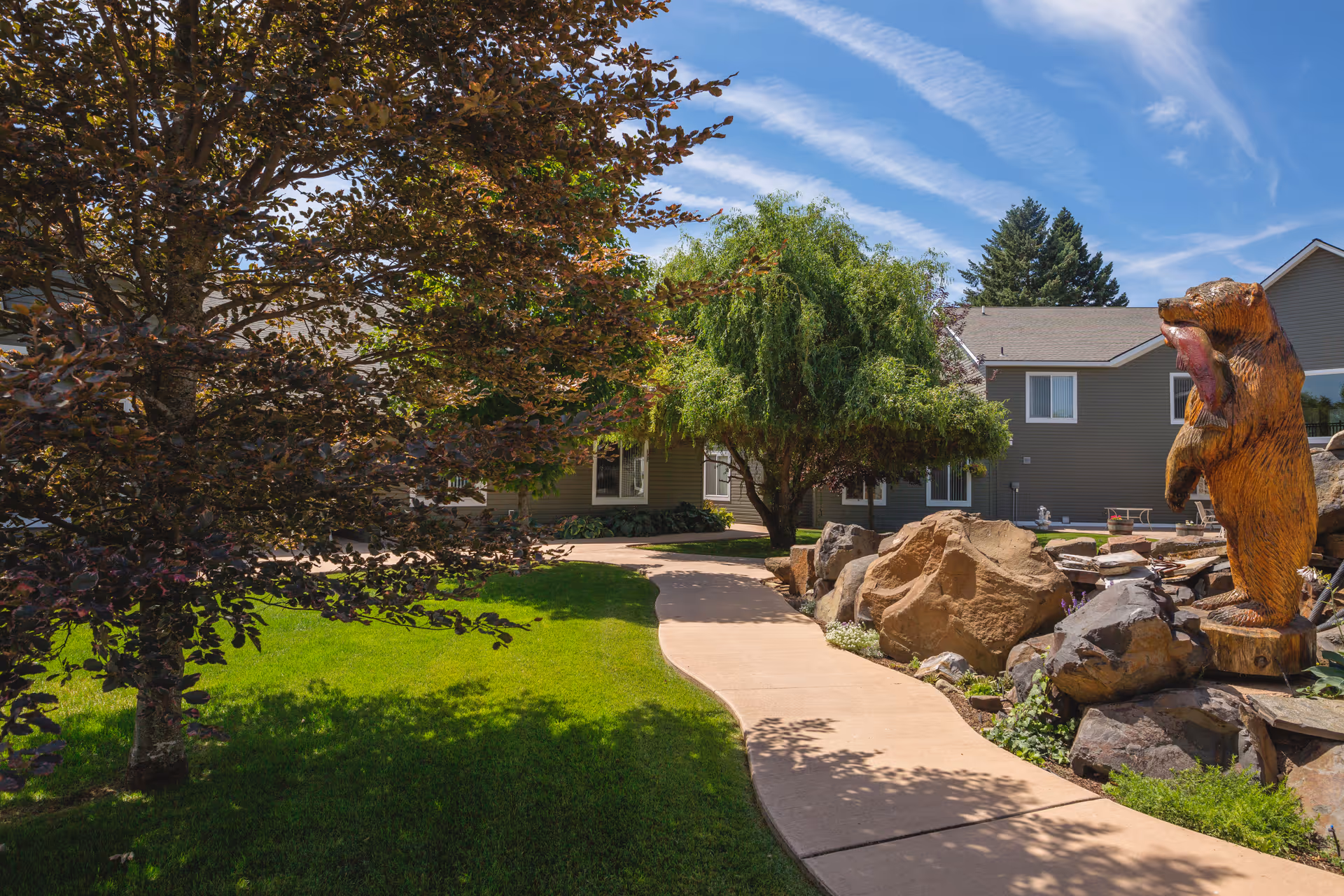 A landscaped outdoor area at Generations Memory Care featuring a curved concrete pathway surrounded by green grass, trees, large rocks, and a wooden bear sculpture holding a fish. The building is visible in the background under a partly cloudy blue sky.