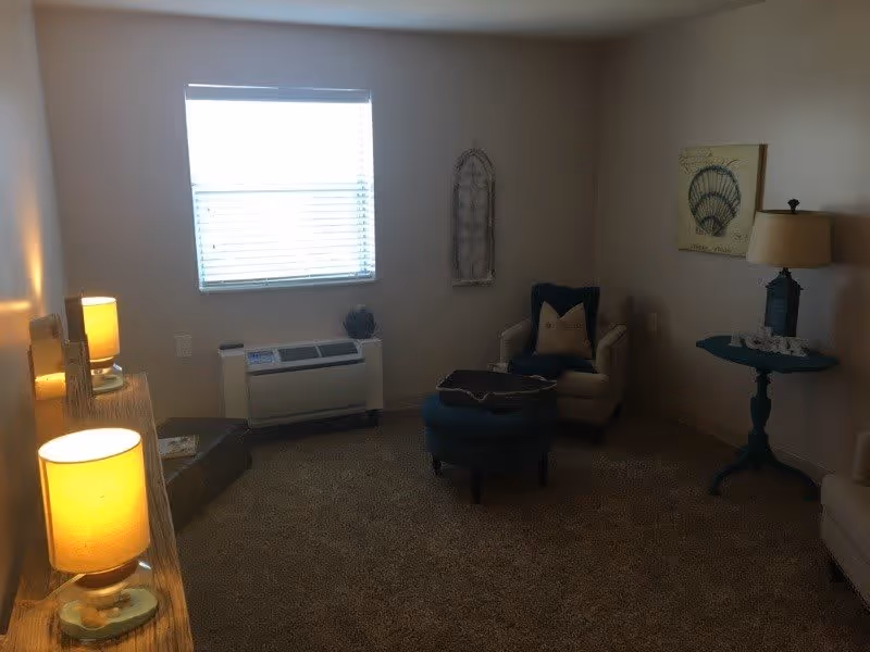 A cozy living room with beige walls and carpet. There is a window with blinds letting in natural light. The room contains a white armchair with a pillow, a blue ottoman, a small round blue table with a lamp and decorative items, and a wooden shelf with two lit lamps. Wall decorations include a framed picture of a seashell and a decorative metal piece.