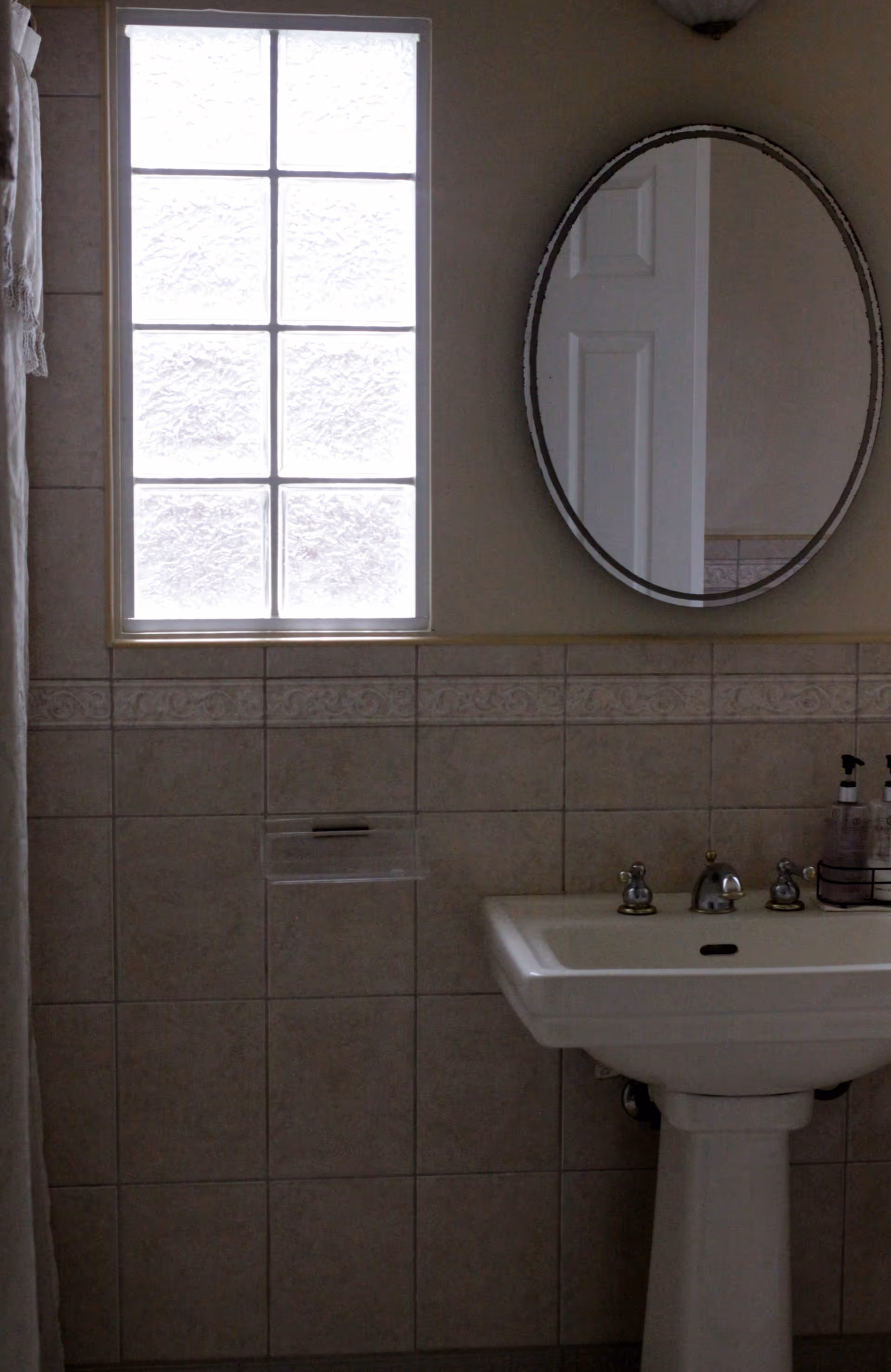 A bathroom interior featuring a frosted glass window with six panes, a round mirror mounted on the wall, and a white pedestal sink with silver faucets. There are soap dispensers on the right side of the sink and beige tiled walls with a decorative border.