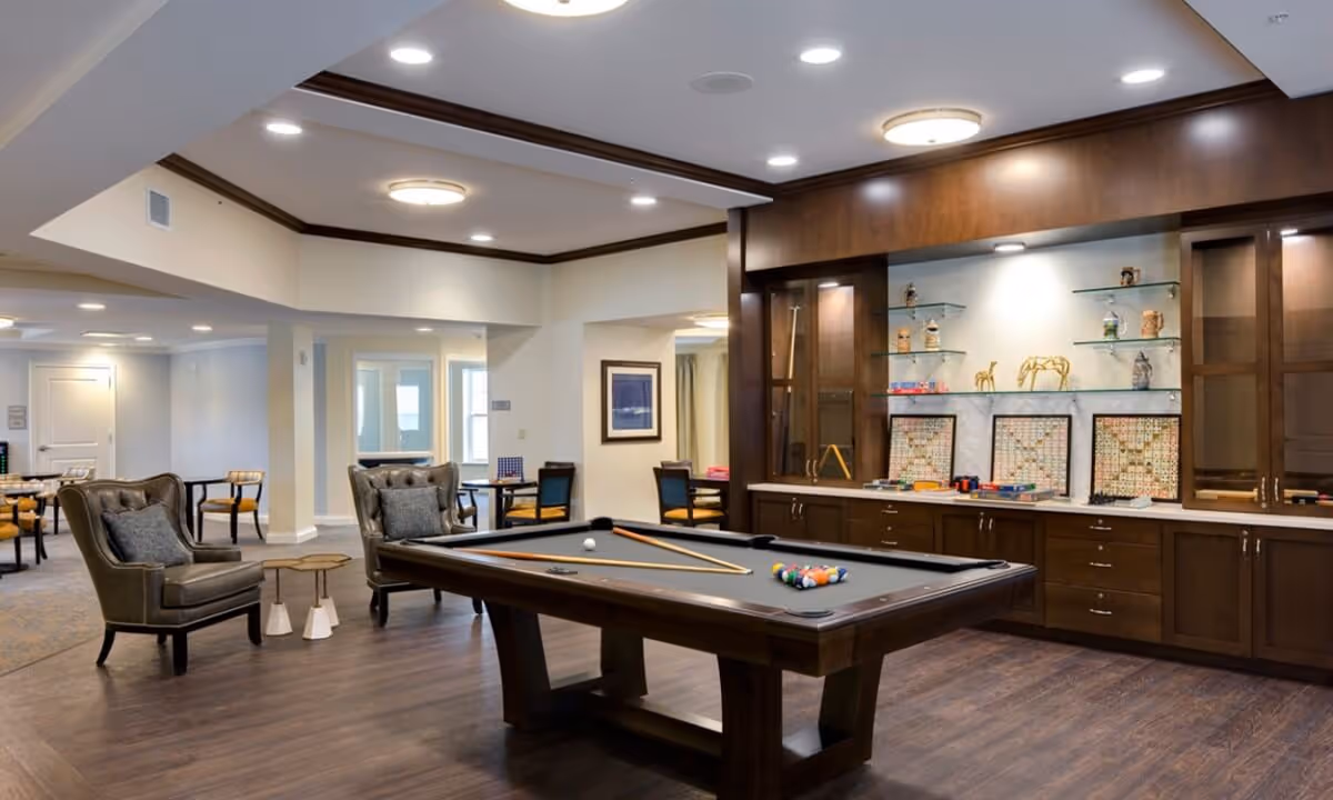 A spacious recreational room with a pool table in the center, two leather armchairs with cushions, and several tables with chairs in the background. The room features wood flooring, recessed lighting, and a built-in wooden cabinet with shelves displaying decorative items and board games.