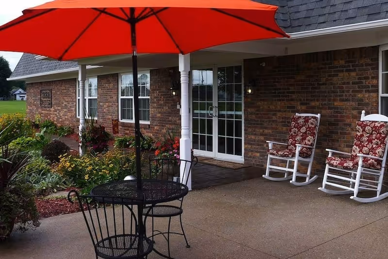 Outdoor patio area with a black metal table and chairs under a large red umbrella, two white rocking chairs with red floral cushions, a brick building with glass double doors, and a garden with various plants and flowers.