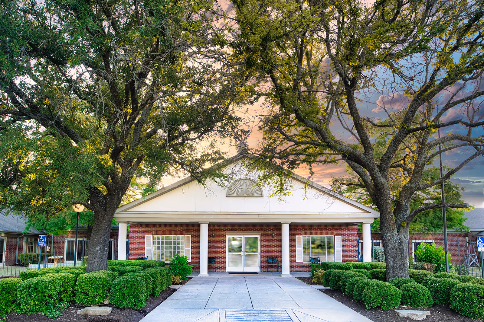Front entrance of a single-story brick nursing facility with white columns, large trees, and a paved walkway.